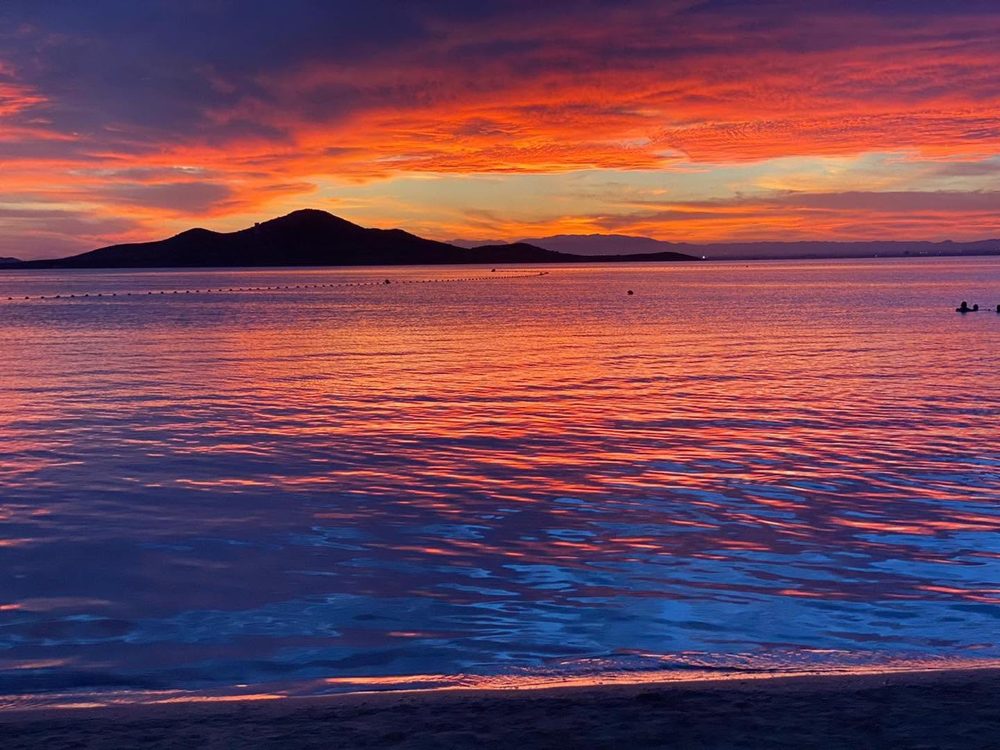 Cielo naranja y morado sobre la silueta de la isla, reflejado en el agua quieta