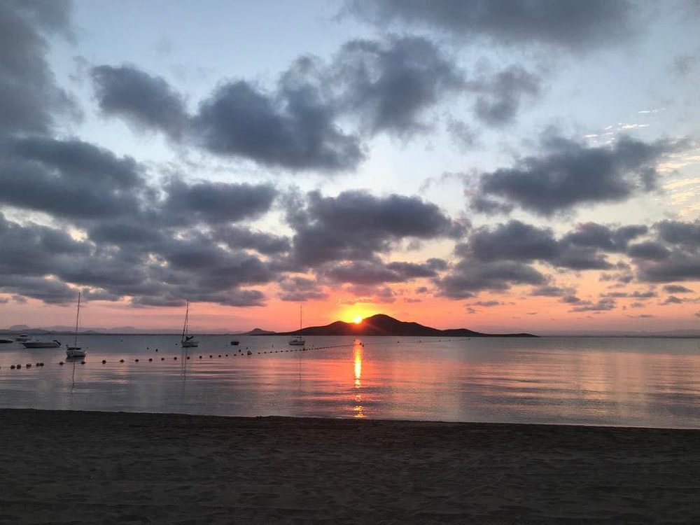 El sol poniéndose justo detrás de la isla, con veleros fondeados y la arena en primer término